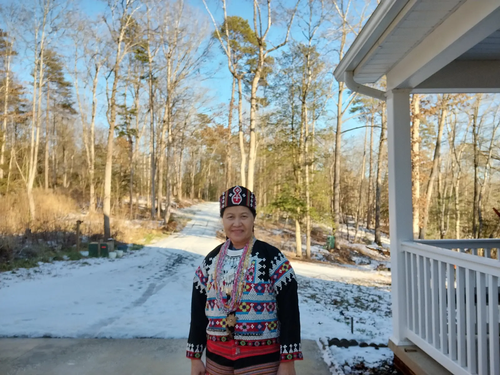 A woman stands outside a house in a snowy wooded area, wearing a traditional Lahu outfit.