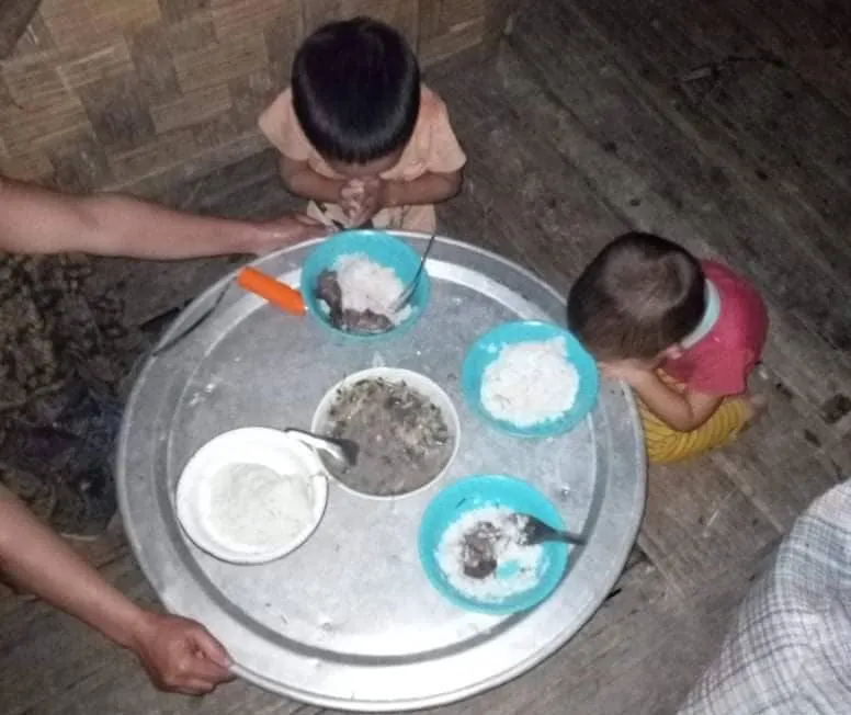 Two children sitting on the ground around a large metal tray with three bowls of food and two spoons.